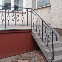 ART-REMONT Robert Skrzypczak - Wykończone schody wejściowe z szarą terakotą i czarną metalową balustradą o wzorze rombów, widoczne ocieplenie cokołu budynku w dwóch odcieniach brązu i biała strukturalna elewacja.