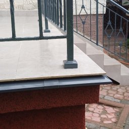 ART-REMONT Robert Skrzypczak - Wykończony taras z beżowymi płytkami, czarną balustradą i schodami, widok na ceglaną ścieżkę i ogród w tle.