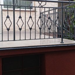 ART-REMONT Robert Skrzypczak - Zadaszony taras z czarną, metalową balustradą z motywem rombów i jasną, ceramiczną podłogą w Warce, widok z poziomu gruntu.