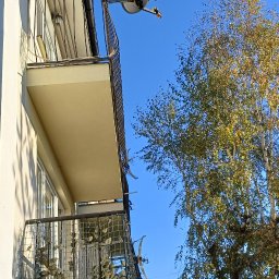 ART-REMONT Robert Skrzypczak - Wysoki budynek z balkonami z metalową siatką, widok z dołu na tle błękitnego nieba i drzew z żółknącymi liśćmi.