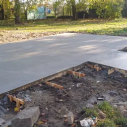 Świeżo wylana, gładka betonowa płyta fundamentowa z drewnianym szalunkiem, widok z poziomu gruntu na tle trawy i drzew.