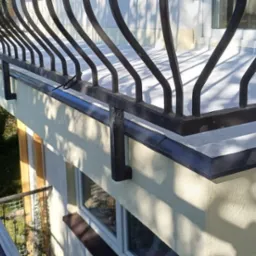 Ocieplona elewacja budynku z balkonem z kutą, czarną balustradą, widok z góry z rusztowania, z widocznym pojemnikiem z zaprawą.