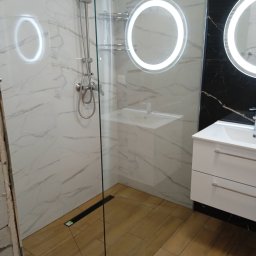 ART-REMONT Robert Skrzypczak - Nowoczesna łazienka z kabiną prysznicową typu walk-in, wykończona marmurowymi płytkami na ścianach i drewnopodobną podłogą, z okrągłymi lustrami podświetlanymi LED i białą szafką pod umywalką.