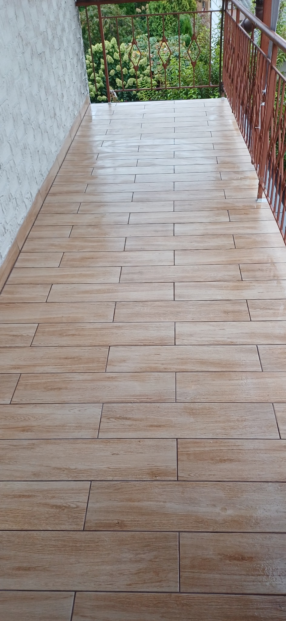 Balkon wyłożony podłużnymi płytkami imitującymi drewno, z metalową balustradą i widokiem na zieleń.