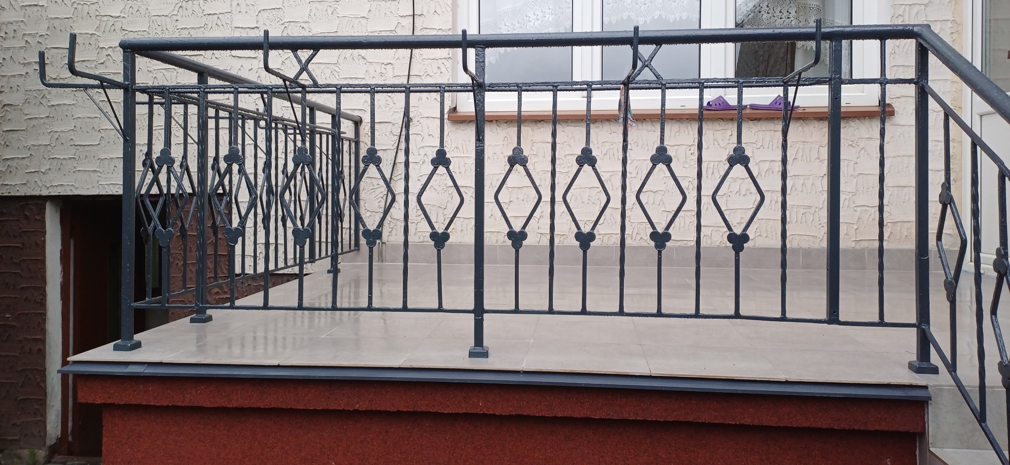 Metalowa balustrada balkonowa w kolorze grafitowym z ozdobnymi elementami w kształcie rombów i czterolistnych koniczynek, widok z dołu na elewację budynku z oknem i parapetem, na którym leżą...