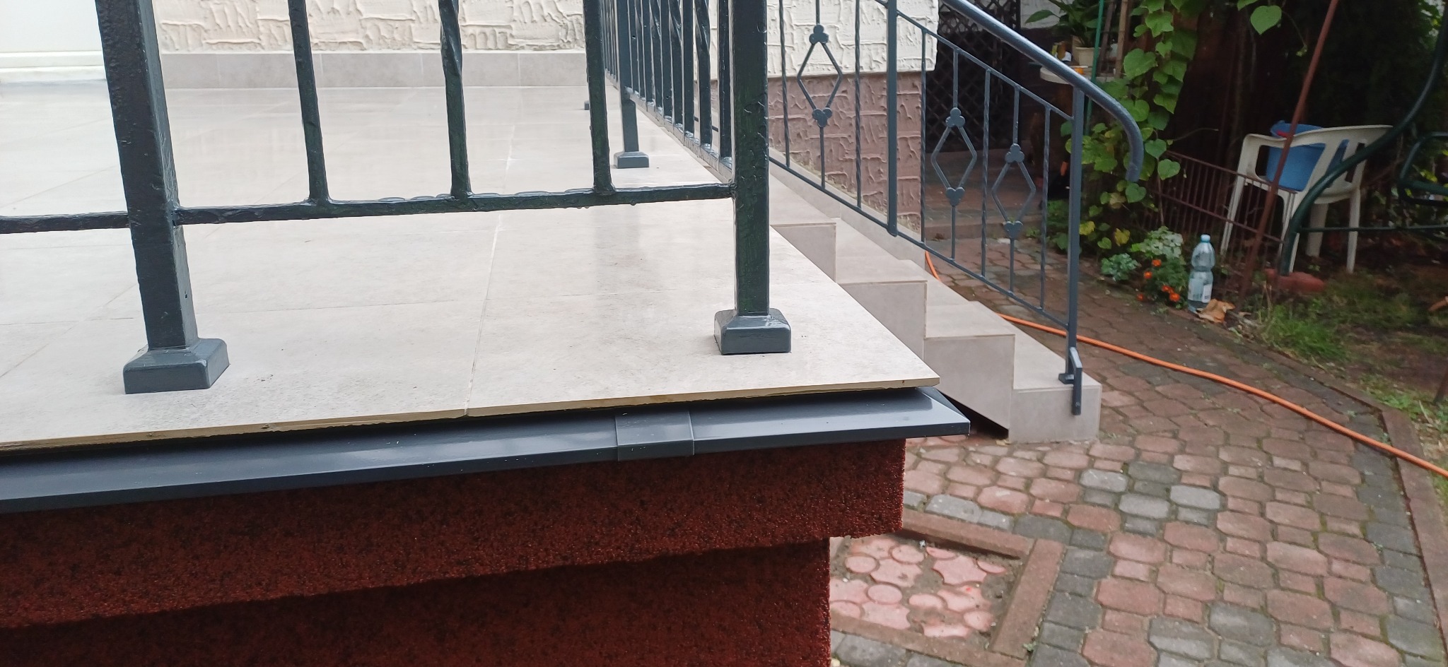 Wykończony taras z beżowymi płytkami, czarną balustradą i schodami, widok na ceglaną ścieżkę i ogród w tle.