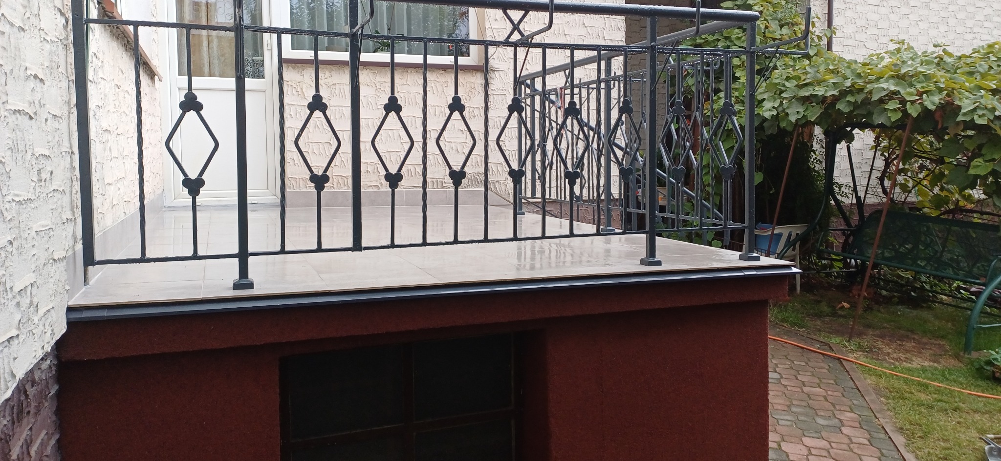 Zadaszony taras z czarną, metalową balustradą z motywem rombów i jasną, ceramiczną podłogą w Warce, widok z poziomu gruntu.