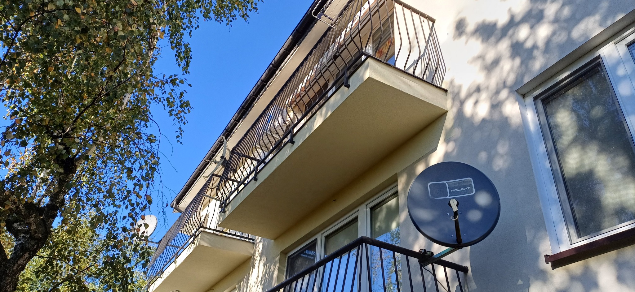 Budynek z dwoma balkonami z czarną, ozdobną balustradą, antena satelitarna na ścianie, widok z dołu na tle błękitnego nieba i drzewa z zielonymi liśćmi.