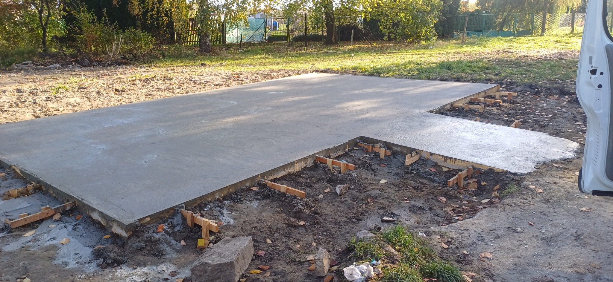 Świeżo wylana, gładka betonowa płyta fundamentowa z drewnianym szalunkiem, widok z poziomu gruntu na tle trawy i drzew.