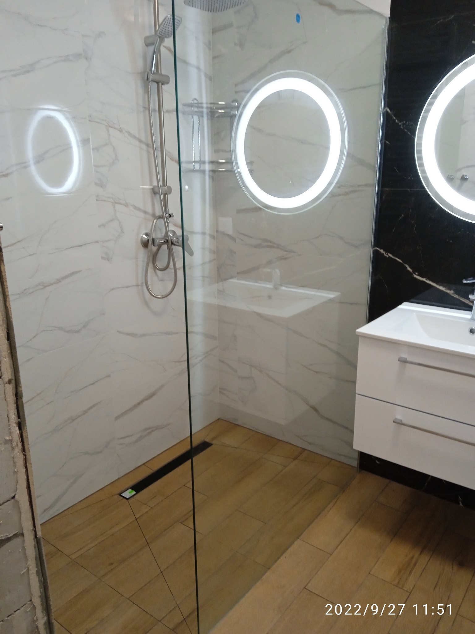 Nowoczesna łazienka z kabiną prysznicową typu walk-in, wykończona marmurowymi płytkami na ścianach i drewnopodobną podłogą, z okrągłymi lustrami podświetlanymi LED i białą szafką pod umywalką.