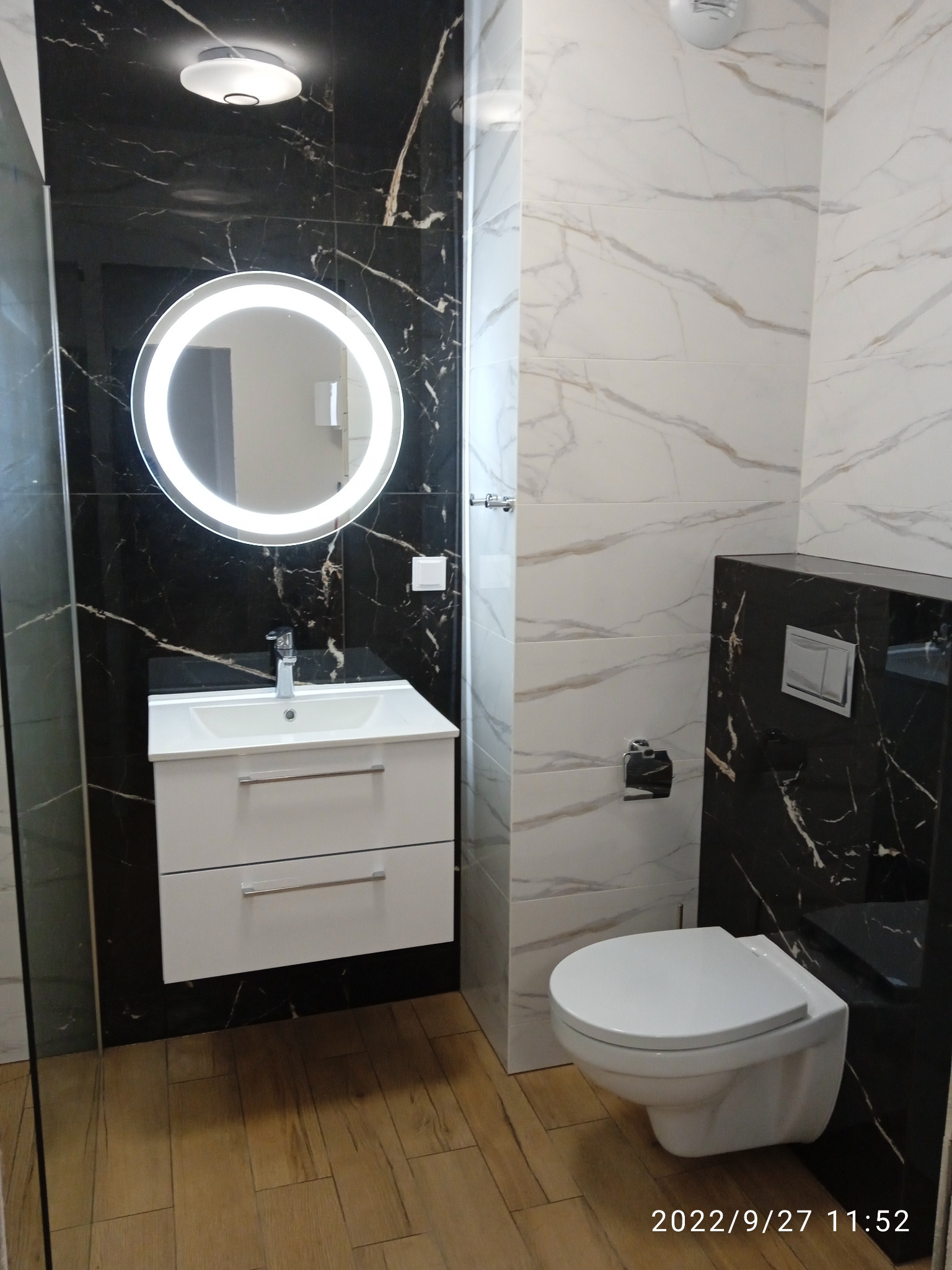 Nowoczesna łazienka z czarno-białymi marmurkowymi płytkami, okrągłym lustrem z podświetleniem LED, białą szafką z umywalką i podwieszaną toaletą.