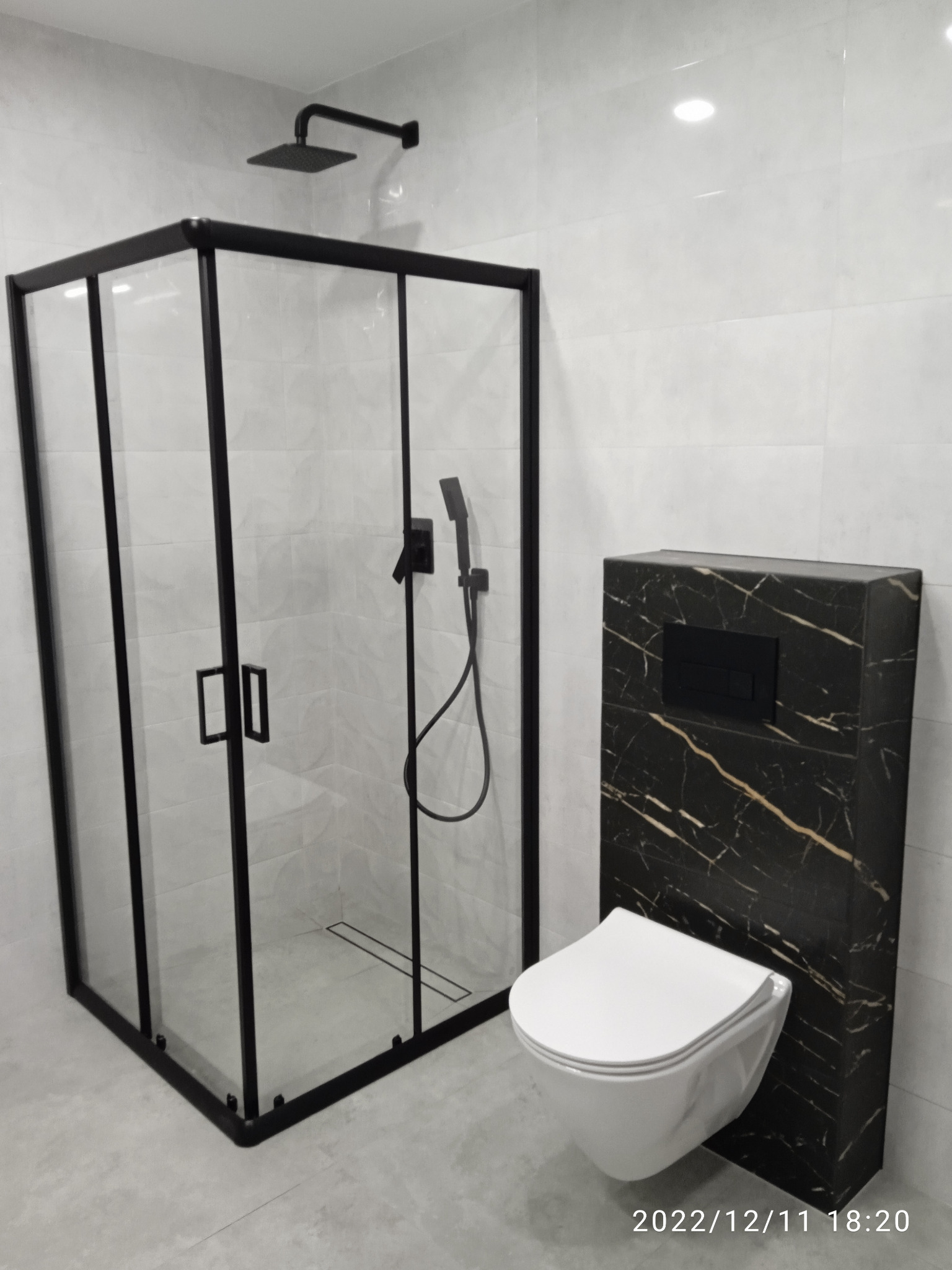 Nowoczesna łazienka z kabiną prysznicową typu walk-in z czarnymi profilami, wiszącą toaletą i zabudową stelaża WC z płyty imitującej czarny marmur z jasnymi żyłami.