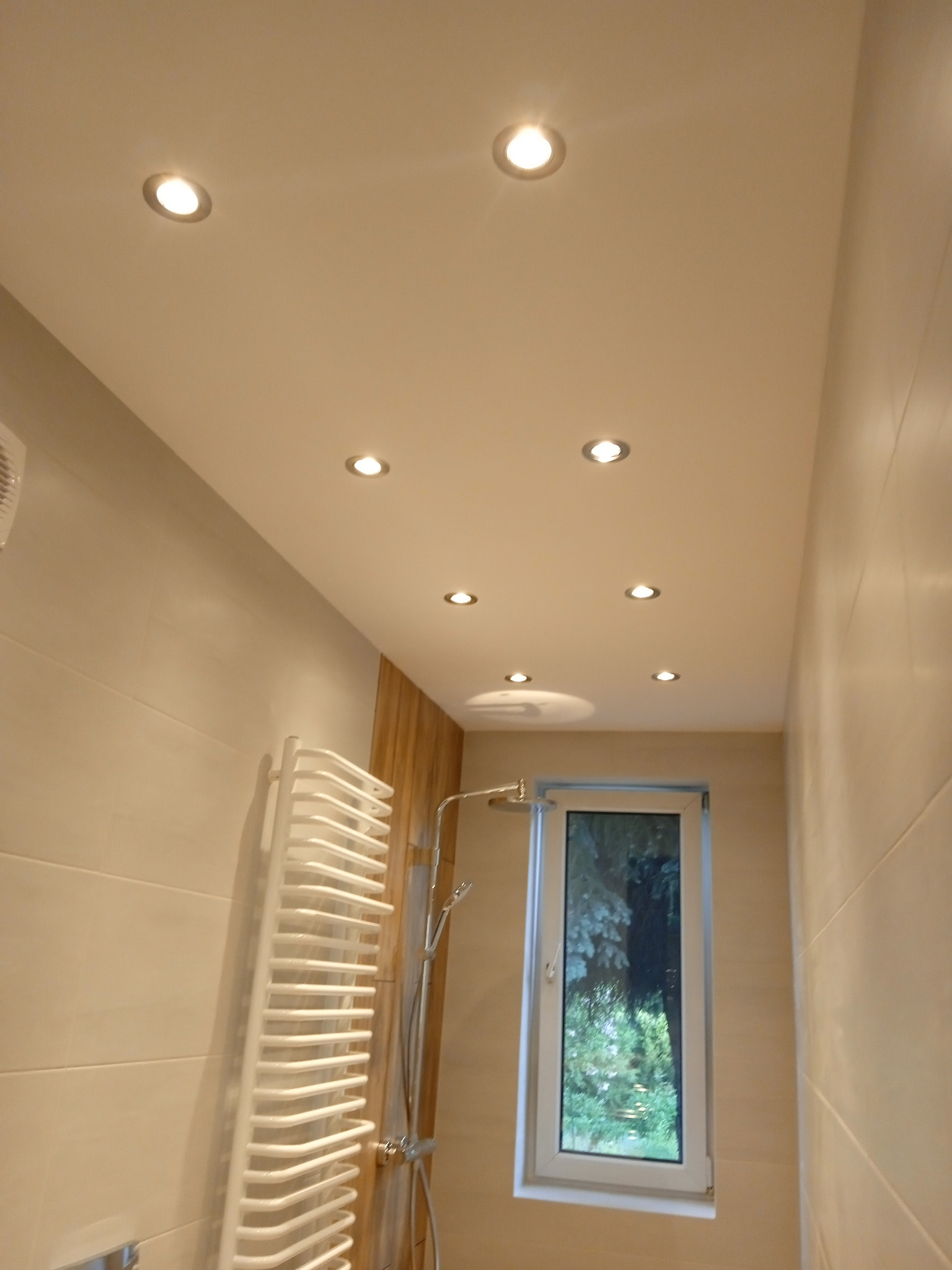 Jasna łazienka z beżowymi płytkami, podwieszanym sufitem z punktowym oświetleniem, grzejnikiem drabinkowym i oknem z widokiem na zieleń.