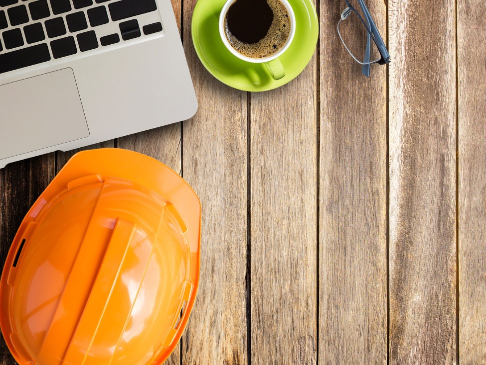 Pomarańczowy kask budowlany, laptop, okulary i filiżanka kawy na drewnianym biurku, widok z góry.
