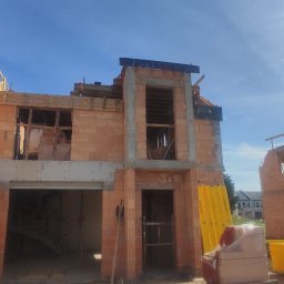 RavBud - Wznoszenie dwukondygnacyjnego budynku z czerwonej cegły ceramicznej, widoczne ściany w stanie surowym otwartym, drewniane szalunki nad otworami okiennymi, materiały budowlane składowane obok.