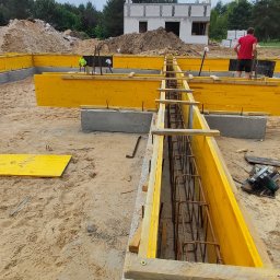 RavBud - Przygotowanie szalunków i zbrojenia pod fundament płyty, widoczne żółte deski szalunkowe, stalowe pręty zbrojeniowe wewnątrz, w tle budynek w stanie surowym otwartym i kopce ziemi.