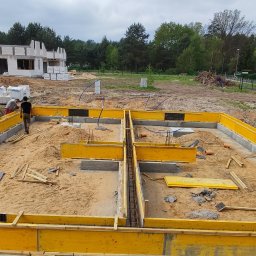 RavBud - Wykopany i częściowo zalany fundament pod budynek z żółtym szalunkiem, widoczne zbrojenia i pracownicy budowlani w tle oraz częściowo wzniesiony budynek z białego bloczka.