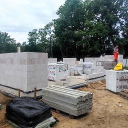 RavBud - Wznoszone ściany z białych bloczków silikatowych na fundamencie, widoczne palety z materiałami budowlanymi Silikat Barlinek i Hydro-Bet Chojnice, pracownik w pomarańczowym kombinezonie...