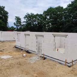 RavBud - Wznoszenie ścian parterowego domu z betonu komórkowego na fundamencie, widoczne otwory okienne i nadproża, w tle drzewa i częściowo ogrodzenie placu budowy.