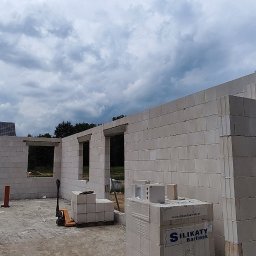 RavBud - Wznoszenie ścian domu z białych bloczków silikatowych, widoczne otwory okienne i drzwiowe, paleta z materiałem budowlanym i pracownik na rusztowaniu w tle.