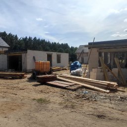 RavBud - Budowa domu z białych bloczków: widok na plac budowy z częściowo wzniesionymi ścianami, drewnianymi szalunkami, materiałami budowlanymi i sprzętem, w tle zalesiony teren i gotowe budynki z ciemną...