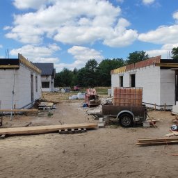 RavBud - Budowa dwóch identycznych budynków z białych bloczków, widoczny szalunek stropu, na pierwszym planie przyczepka z zbiornikiem na wodę, w tle las i błękitne niebo z chmurami.