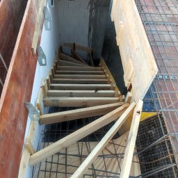 RavBud - Wnętrze domu w trakcie budowy: drewniane szalunki, tymczasowe schody spiralne z surowego drewna prowadzące na wyższą kondygnację, widoczne zbrojenie stropu.