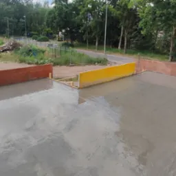 Wylany fundament z widocznym zbrojeniem i szalunkiem z desek w kolorze pomarańczowym i żółtym, z odbiciem nieba w mokrym betonie.