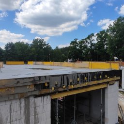 RavBud - Wylewanie betonowego stropu na budowie domu z bloczków, widoczne szalunki i zbrojenia, w tle błękitne niebo z chmurami i zieleń drzew.