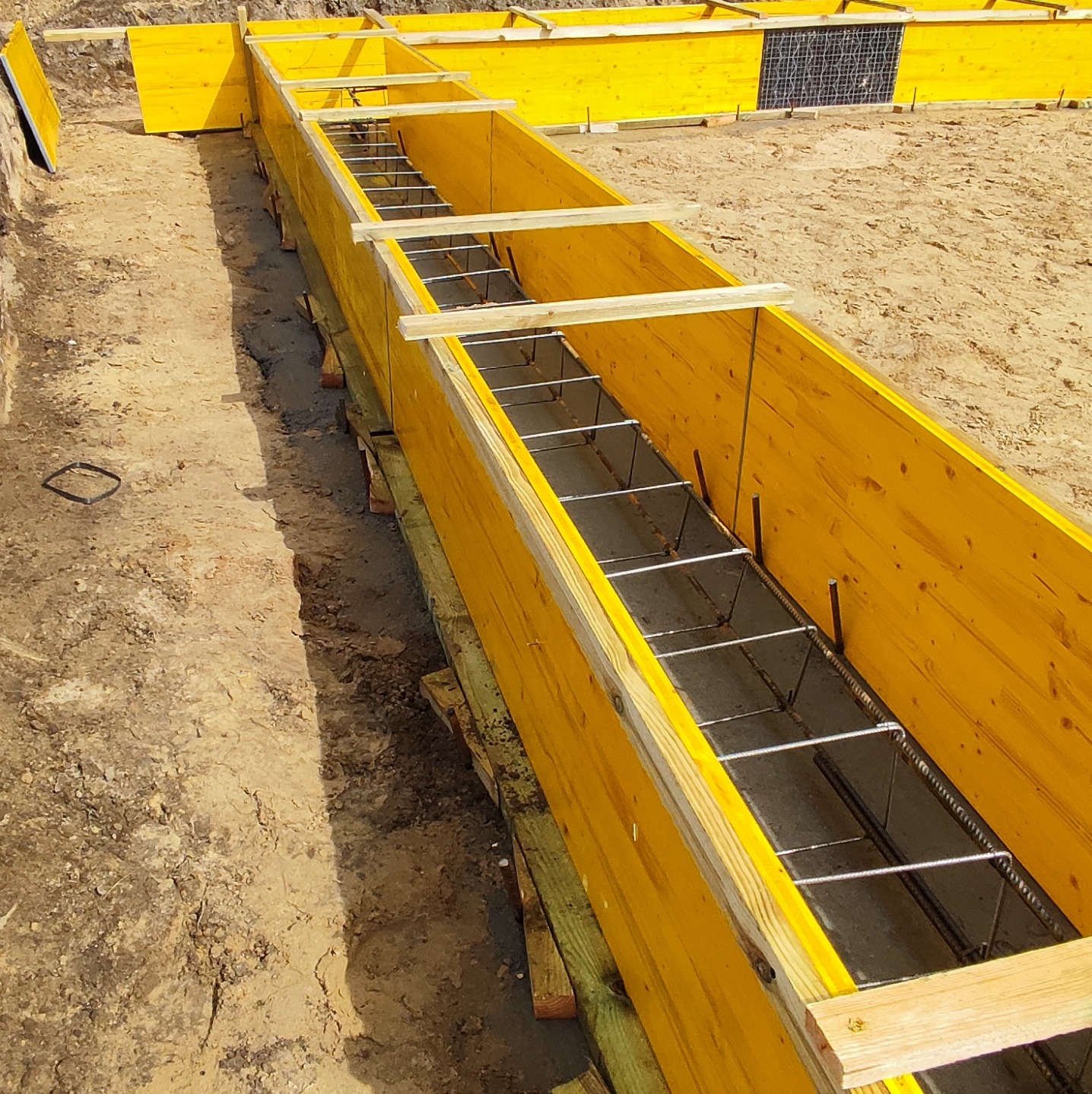 Żółte szalunki fundamentowe zbrojone stalą, przygotowane do wylania betonu, widok z góry na placu budowy.