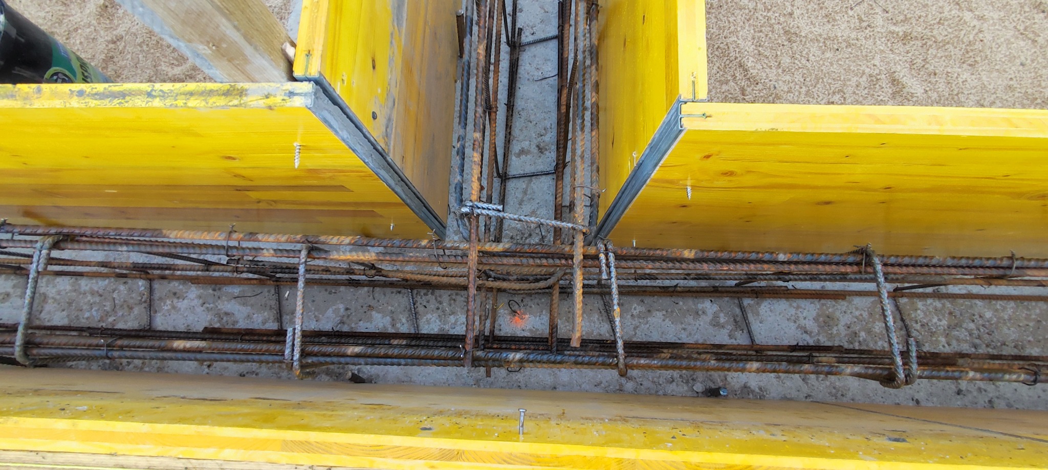 Szczegółowe ujęcie szalunku fundamentowego zbrojonego stalą, widoczne żółte deski szalunkowe i pręty zbrojeniowe połączone drutem.