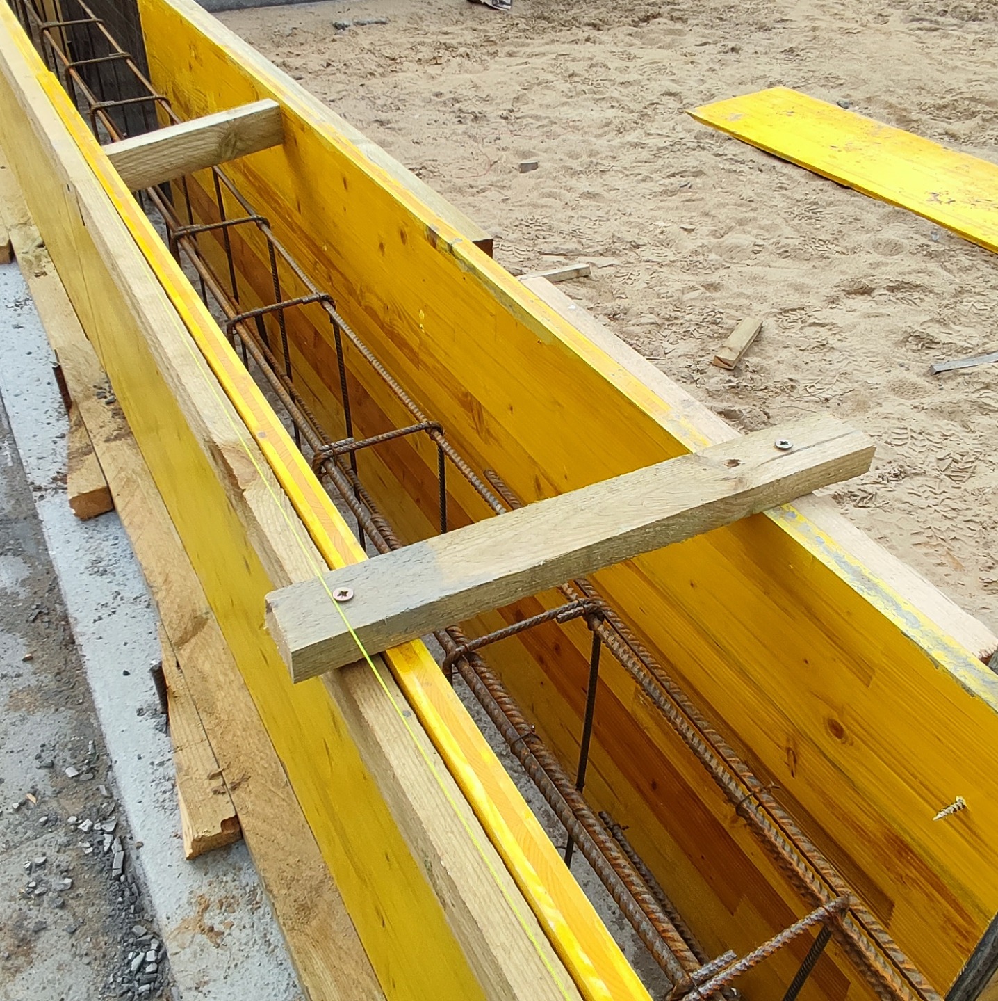 Żółte szalunki zbrojeniowe przygotowane do zalewania fundamentów, widoczne zbrojenie i drewniane podpory, teren budowy w tle.