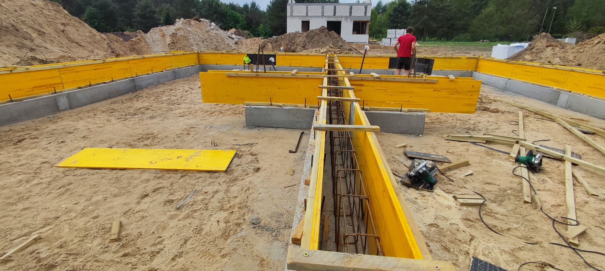Przygotowanie szalunków i zbrojenia pod fundament płyty, widoczne żółte deski szalunkowe, stalowe pręty zbrojeniowe wewnątrz, w tle budynek w stanie surowym otwartym i kopce ziemi.