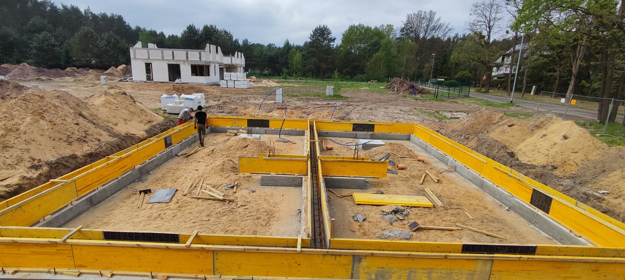 Wykopany i częściowo zalany fundament pod budynek z żółtym szalunkiem, widoczne zbrojenia i pracownicy budowlani w tle oraz częściowo wzniesiony budynek z białego bloczka.