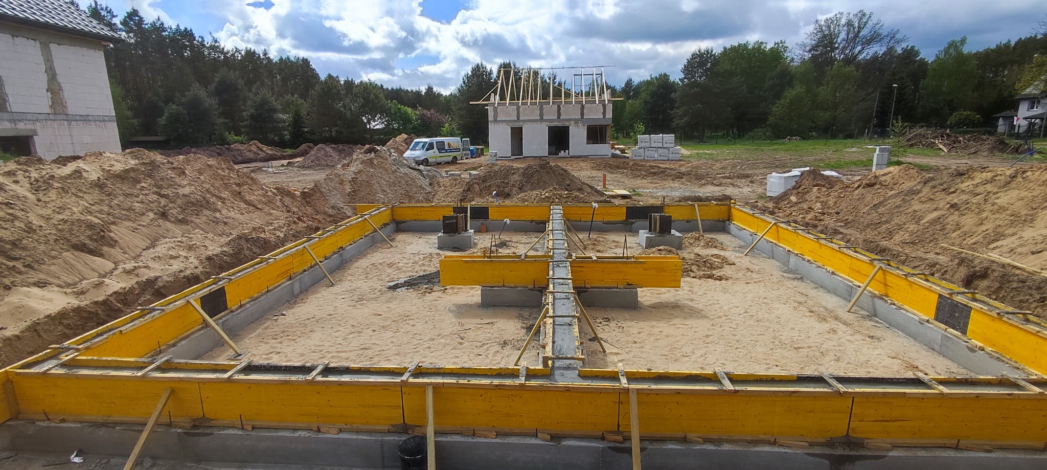 Żółte szalunki fundamentowe na piasku, w tle wznoszony budynek w stanie surowym otwartym i stosy ziemi.