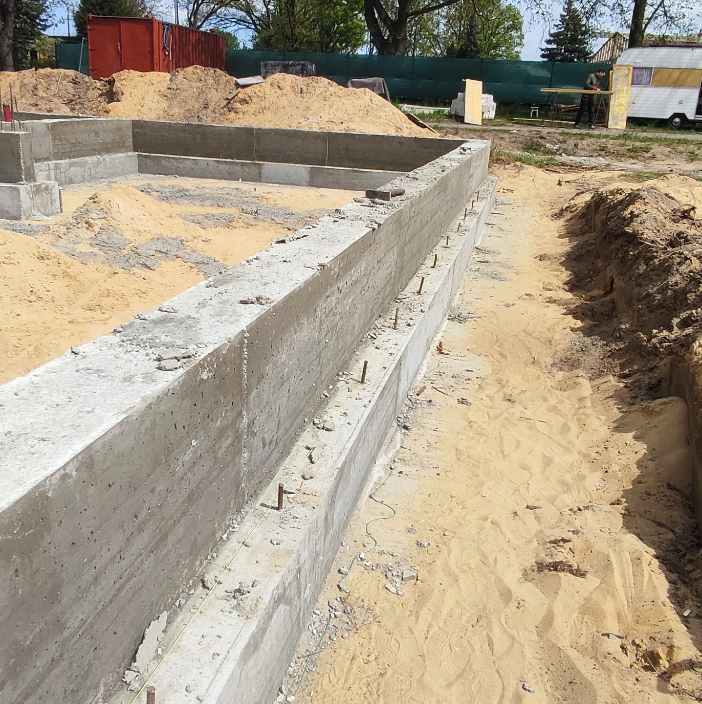 Wylany betonowy fundament z wystającymi prętami zbrojeniowymi, otoczony piaskiem, z widoczną wykopaną linią pod kolejny etap w tle kontener i przyczepa kempingowa.