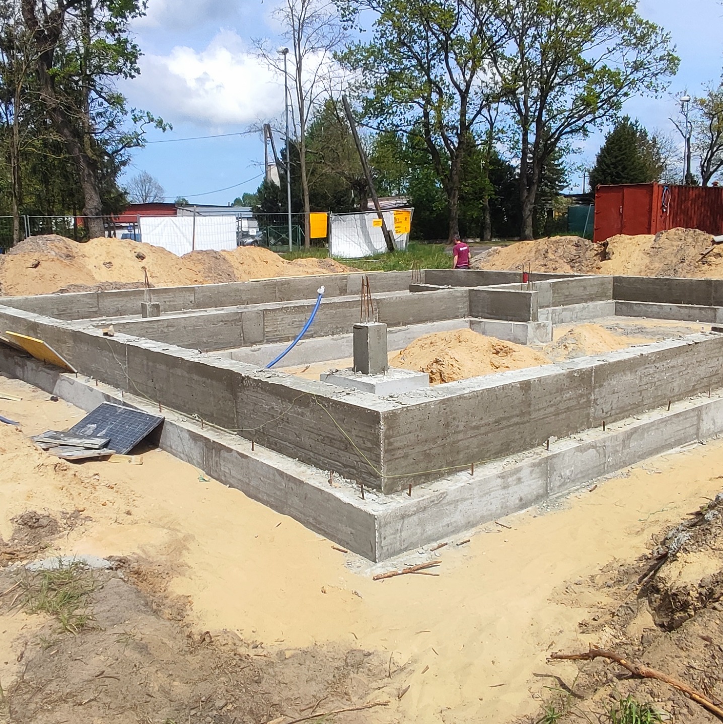 Zalany betonowy fundament domu jednorodzinnego, widoczne wystające pręty zbrojeniowe, piasek wewnątrz fundamentu, w tle drzewa i kontener budowlany.
