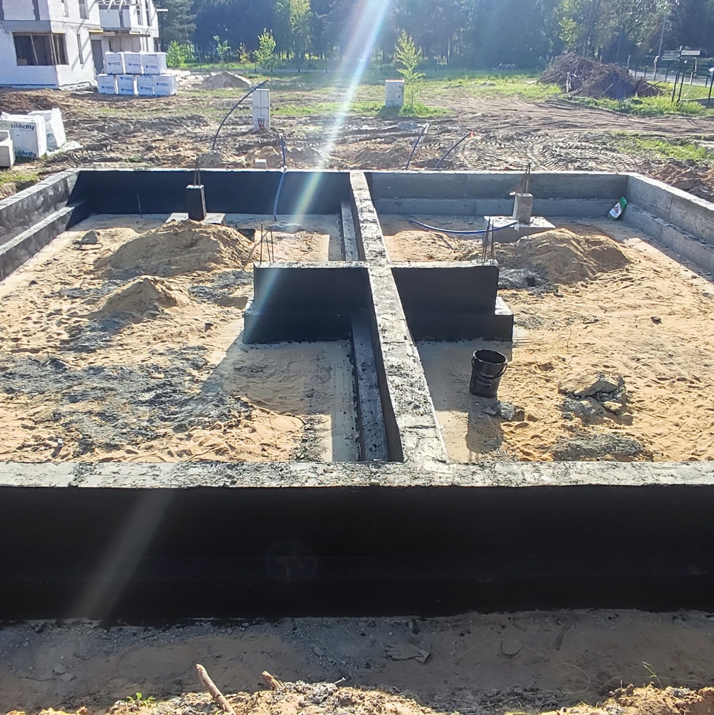 Zalane betonem fundamenty domu z hydroizolacją pionową, widoczny piasek wewnątrz wykopu, w tle palety z bloczkami silikatowymi i budynek w stanie surowym otwartym.