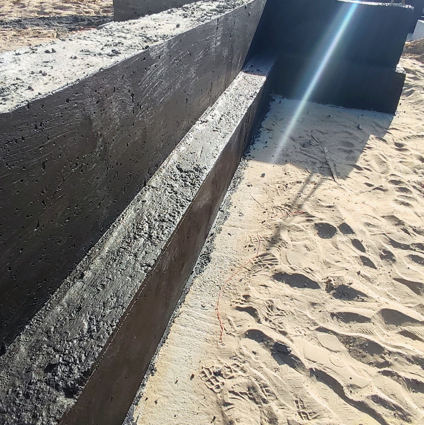 Świeżo wylany, czarny fundament z widocznymi odciskami butów w piasku wokół, z lekkim odblaskiem słońca.