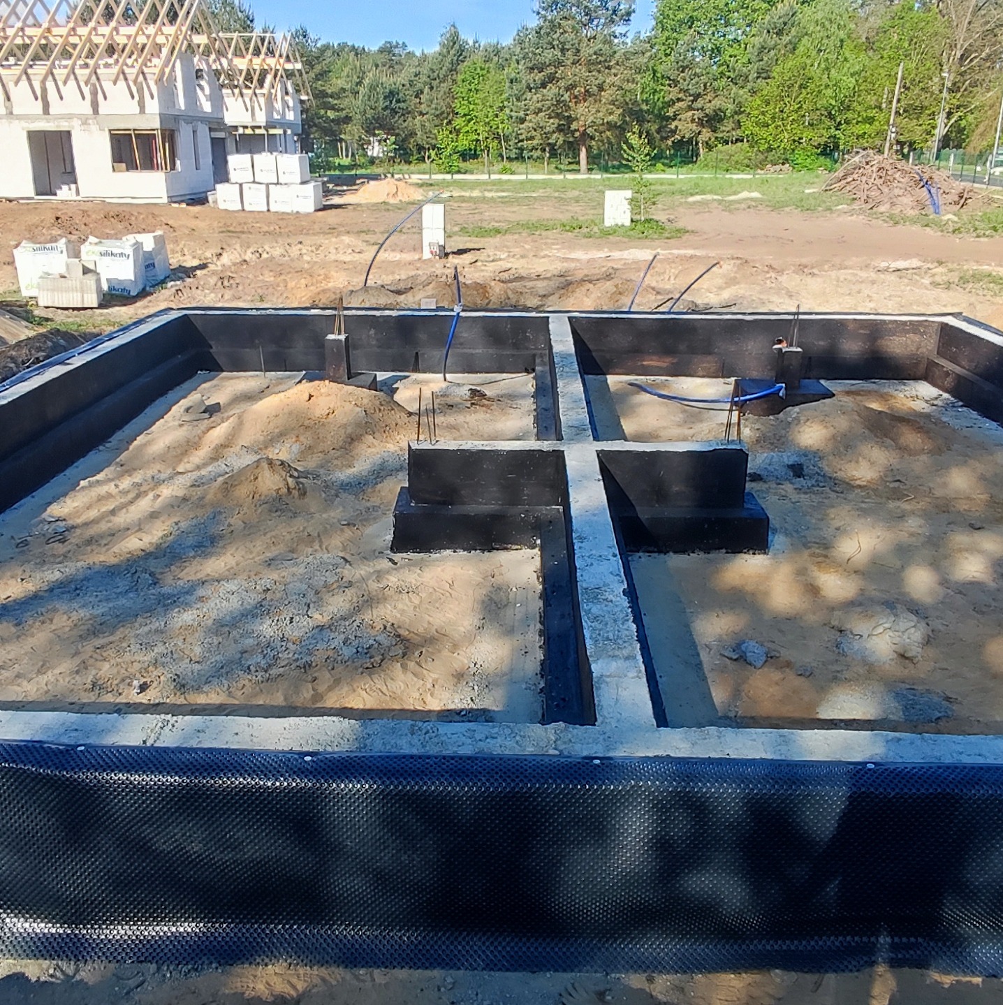Zalany betonem szalunek fundamentowy z izolacją pionową, widoczne wystające pręty zbrojeniowe, w tle budowa domu w stanie surowym otwartym.
