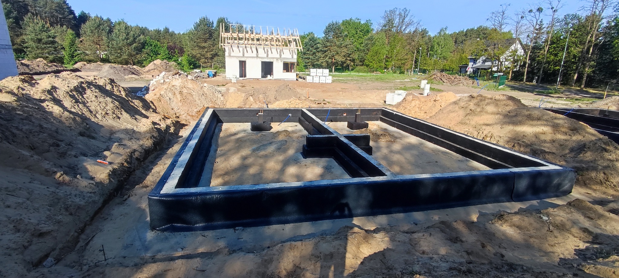Przygotowany fundament pod budowę domu jednorodzinnego z widoczną hydroizolacją, w tle budynek w stanie surowym otwartym z drewnianą konstrukcją dachu.
