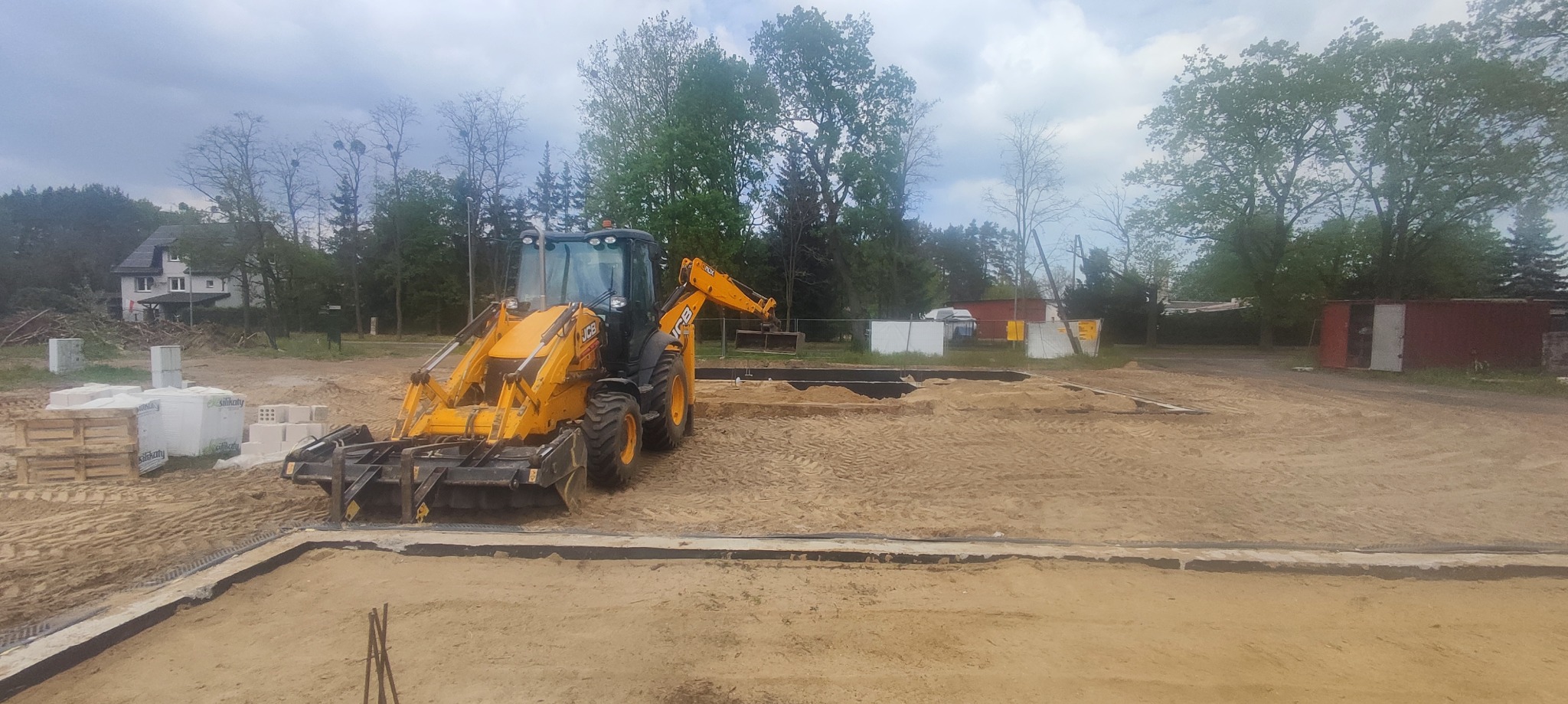 Żółta koparko-ładowarka JCB przy wykopie fundamentów, widoczne bloczki silikatowe na paletach, w tle dom jednorodzinny i drzewa.