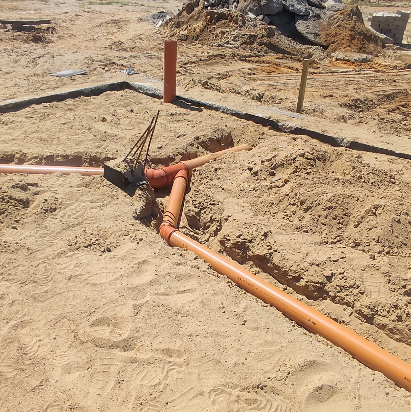Wykonanie instalacji kanalizacyjnej z pomarańczowych rur w wykopie fundamentowym, widoczne zbrojenie i piasek.