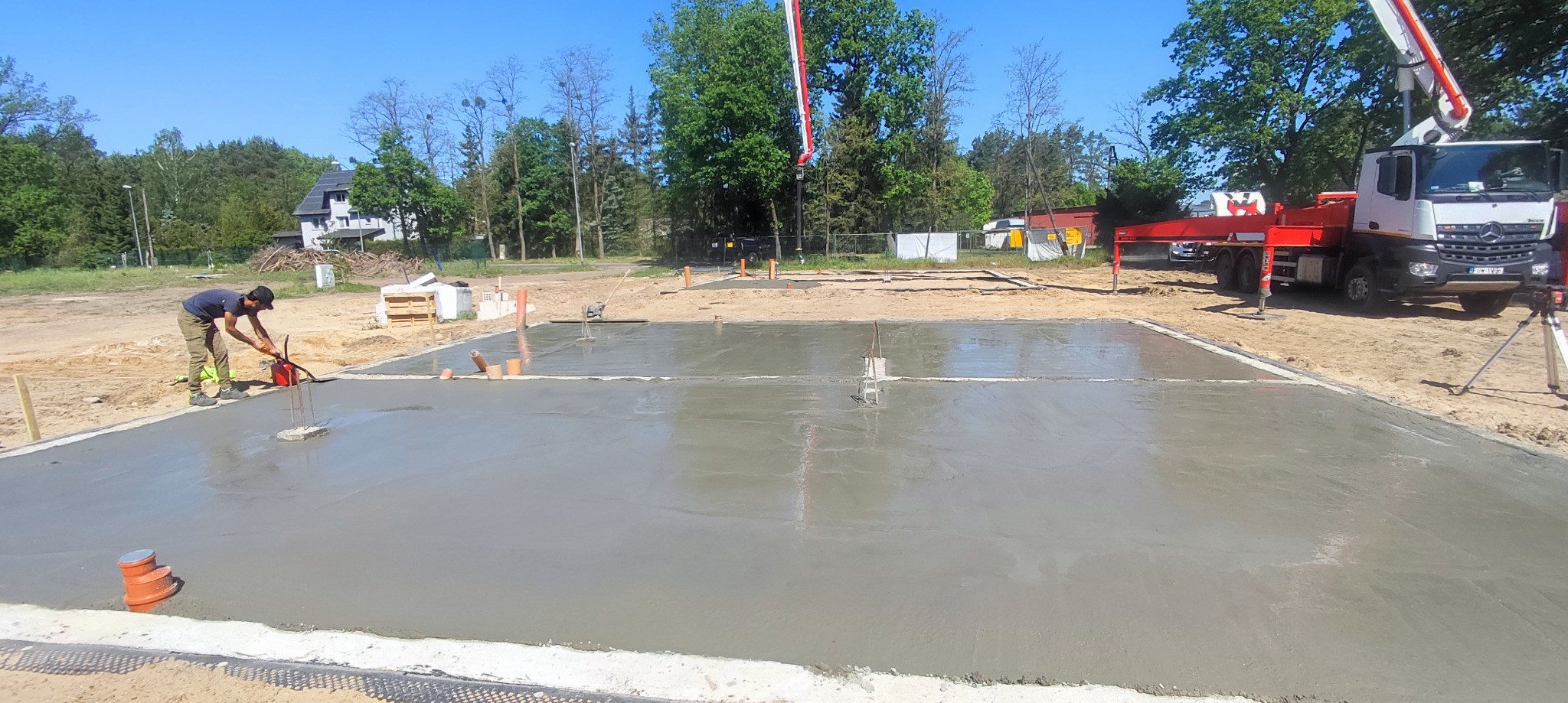 Wylewanie i wyrównywanie betonu na przygotowanym fundamencie pod budowę domu, widoczny pracownik zacieraczką mechaniczną, rury kanalizacyjne, betonowóz z pompą oraz zarys przyszłego budynku.