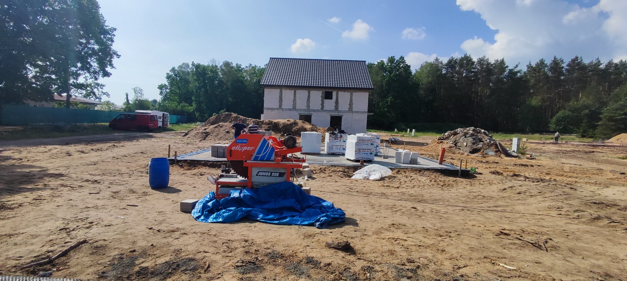 Wczesny etap budowy domu: fundamenty wylane, ściany parteru wzniesione, na pierwszym planie piła do cięcia bloczków z osłoną, w tle las i błękitne niebo.