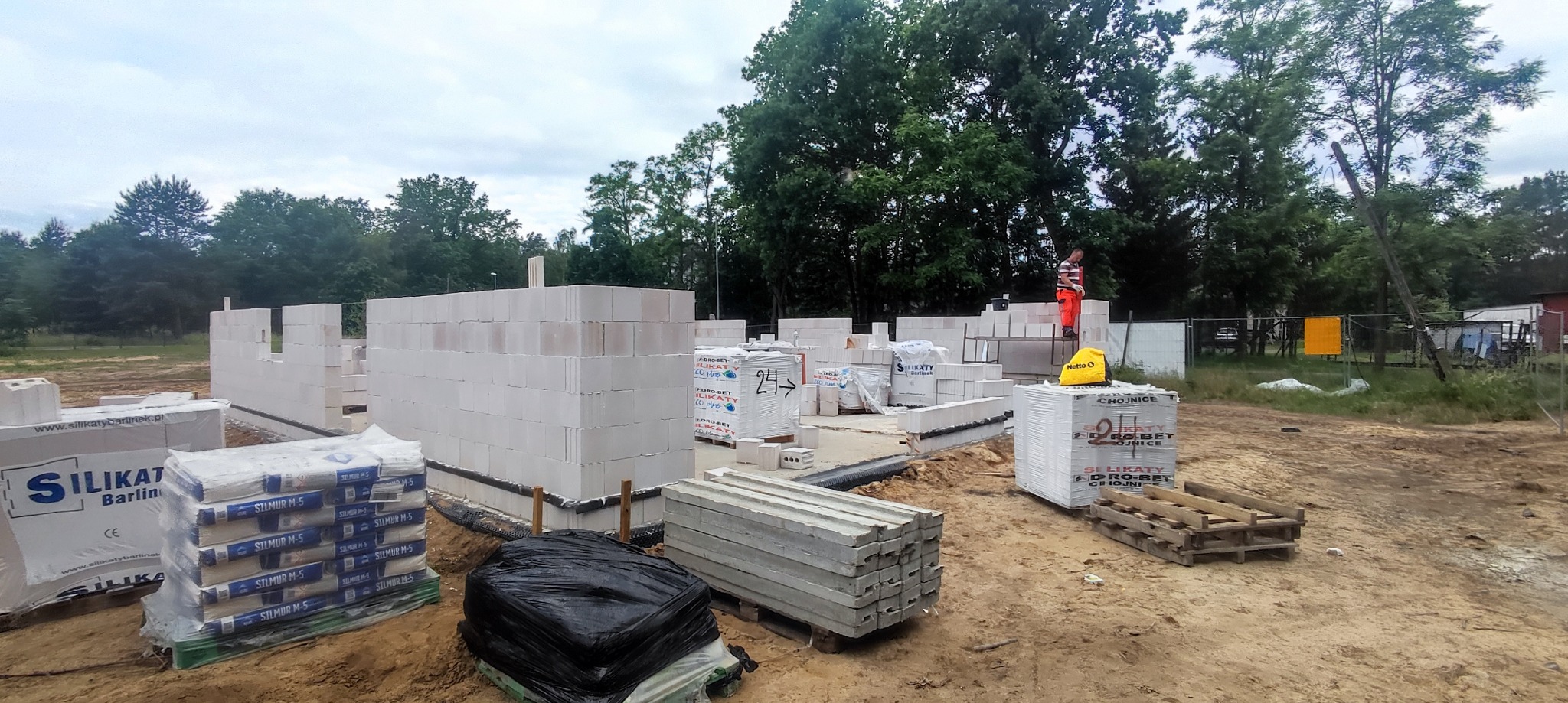 Wznoszone ściany z białych bloczków silikatowych na fundamencie, widoczne palety z materiałami budowlanymi Silikat Barlinek i Hydro-Bet Chojnice, pracownik w pomarańczowym kombinezonie...