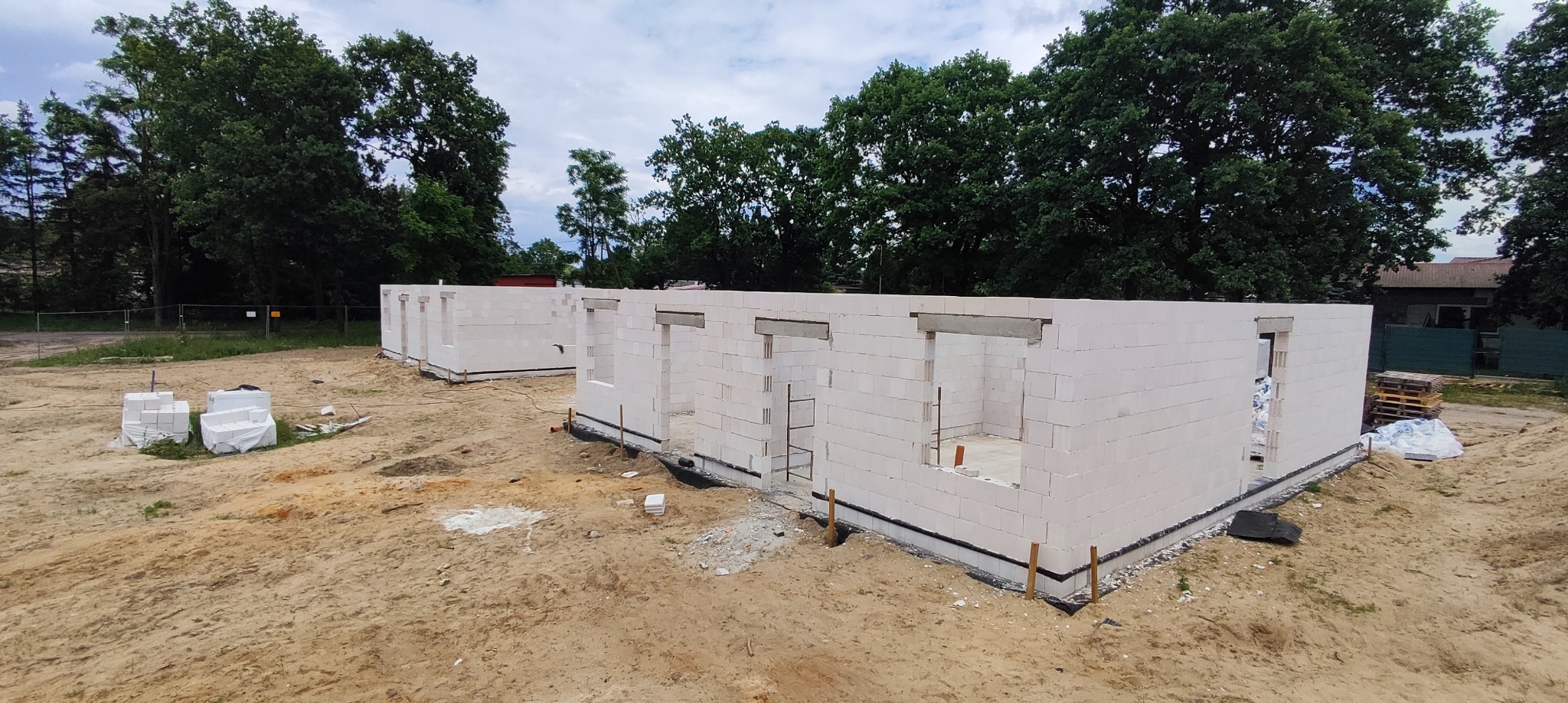 Wznoszenie ścian parterowego domu z betonu komórkowego na fundamencie, widoczne otwory okienne i nadproża, w tle drzewa i częściowo ogrodzenie placu budowy.