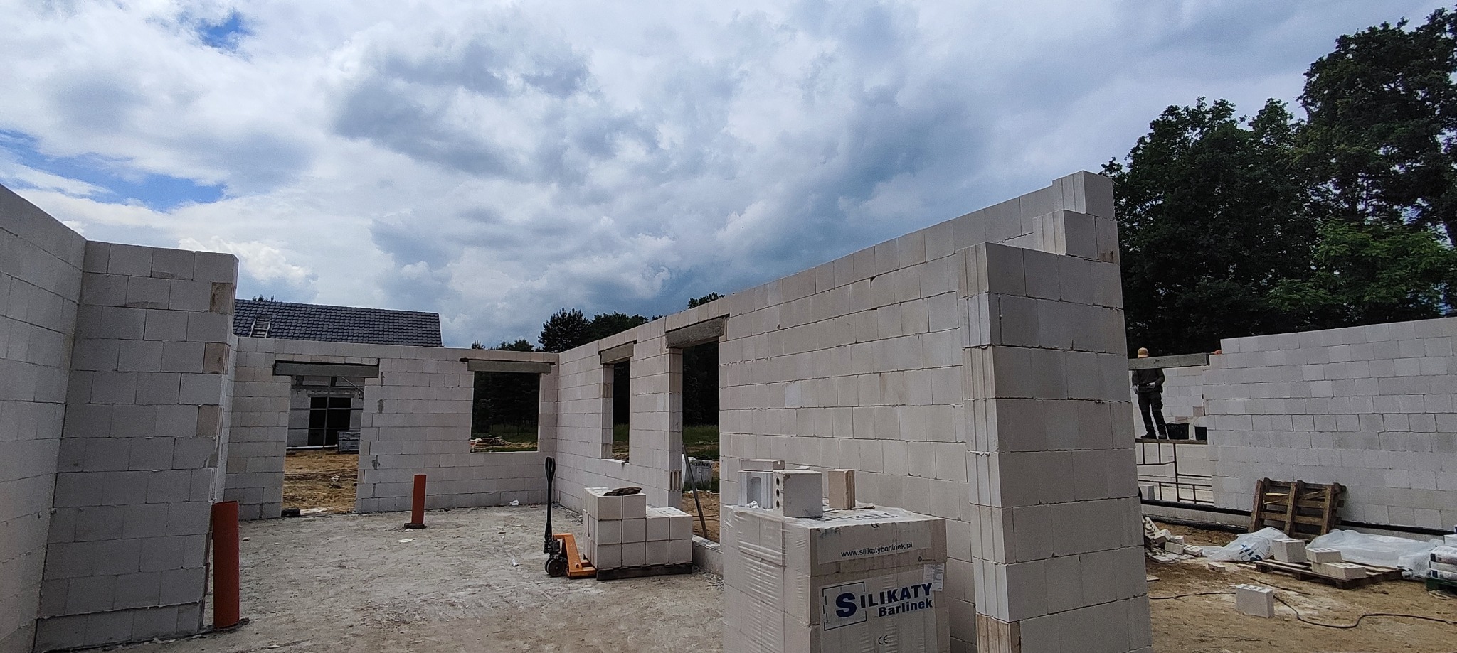 Wznoszenie ścian domu z białych bloczków silikatowych, widoczne otwory okienne i drzwiowe, paleta z materiałem budowlanym i pracownik na rusztowaniu w tle.