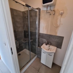 Ewbau - Łazienka w trakcie prac: kabina prysznicowa z ciemnymi płytkami, biała szafka z umywalką, bojler elektryczny na ścianie z widocznymi śladami po instalacji.