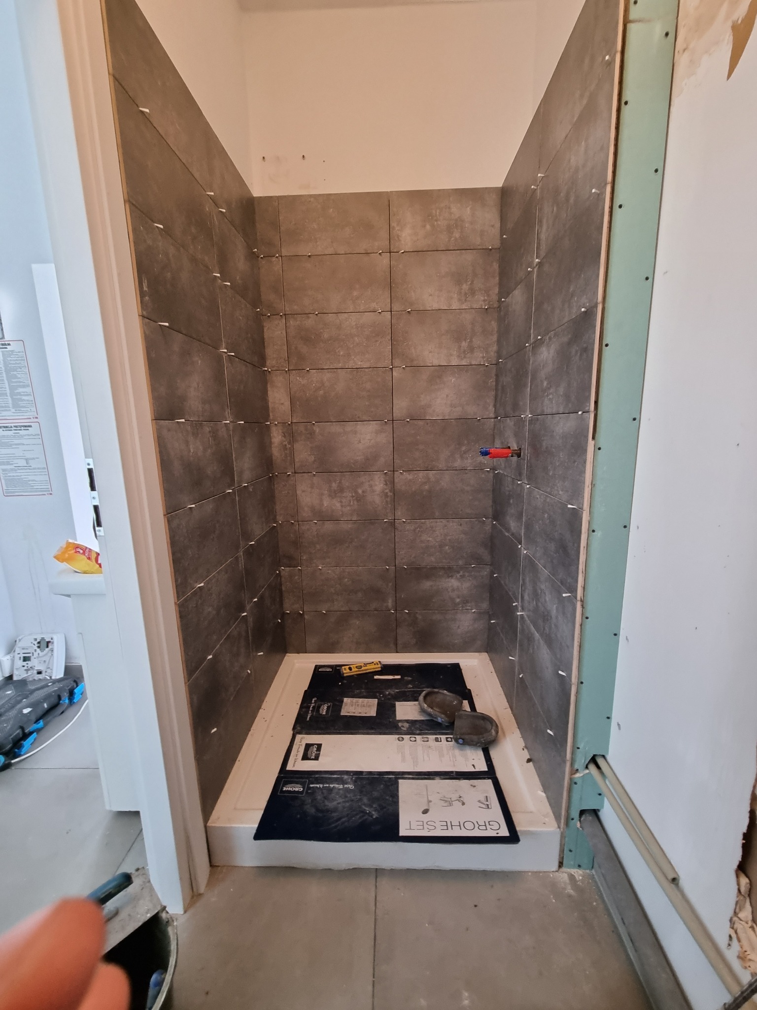 Montaż ciemnych, prostokątnych płytek w kabinie prysznicowej z użyciem krzyżyków dystansowych, widoczne opakowania po zestawie prysznicowym Grohe.